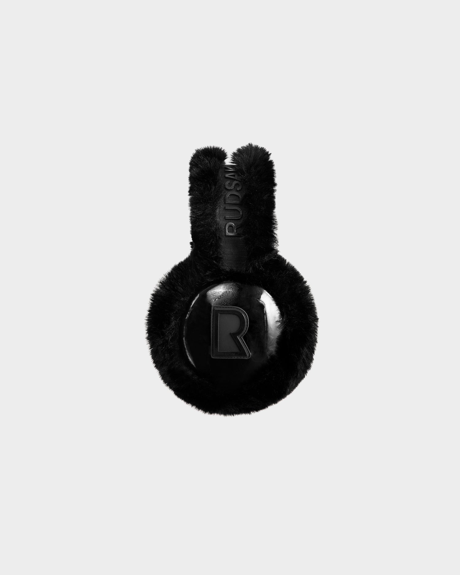 VALENCIA - 8425543 Black by Rudsak: Black faux fur earmuffs with shiny round ear coverings featuring a raised R and "RUSSI" on the headband, displayed on a plain light gray background—a chic accessory for cold weather.