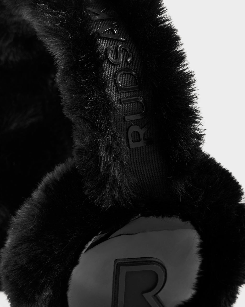 Close-up of VALENCIA - 8425543 Black faux fur earmuffs by Rudsak, featuring the brand name embossed on the headband and a raised R logo on the ear cover, set against a plain light background—a chic winter accessory.