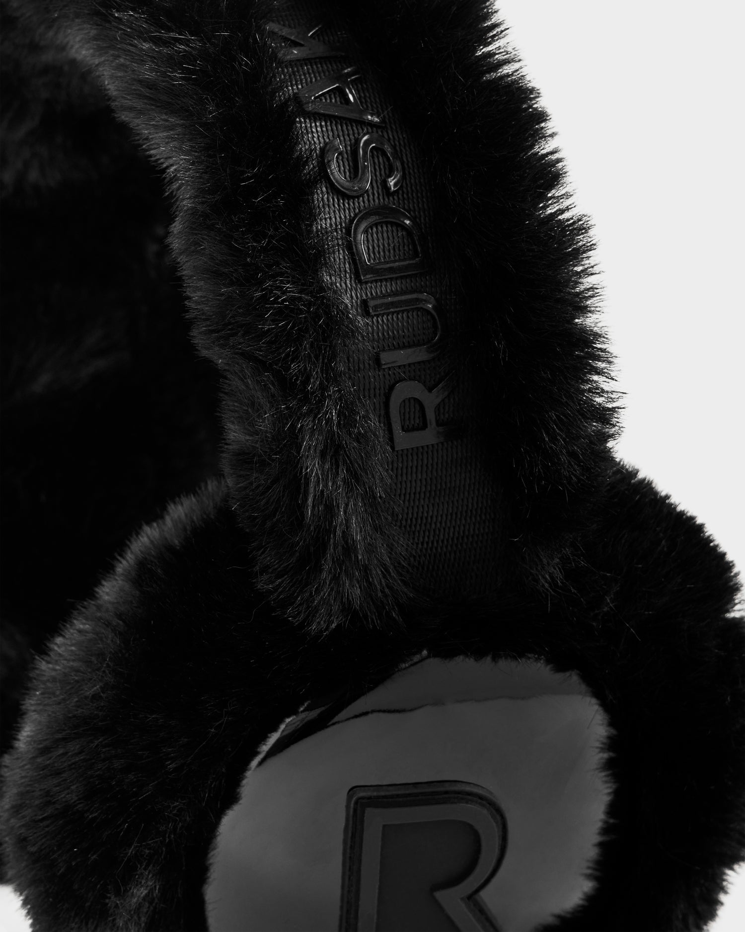 Close-up of VALENCIA - 8425543 Black faux fur earmuffs by Rudsak, featuring the brand name embossed on the headband and a raised R logo on the ear cover, set against a plain light background—a chic winter accessory.