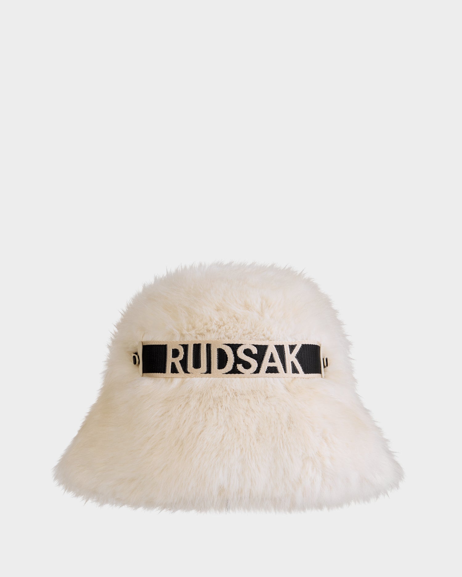 The ROMINA - 8425545 Vanilla by Rudsak is a white faux fur women's bucket hat with a black band featuring bold white RUDSAK lettering, offering cozy-luxe winter style against a simple, light backdrop.