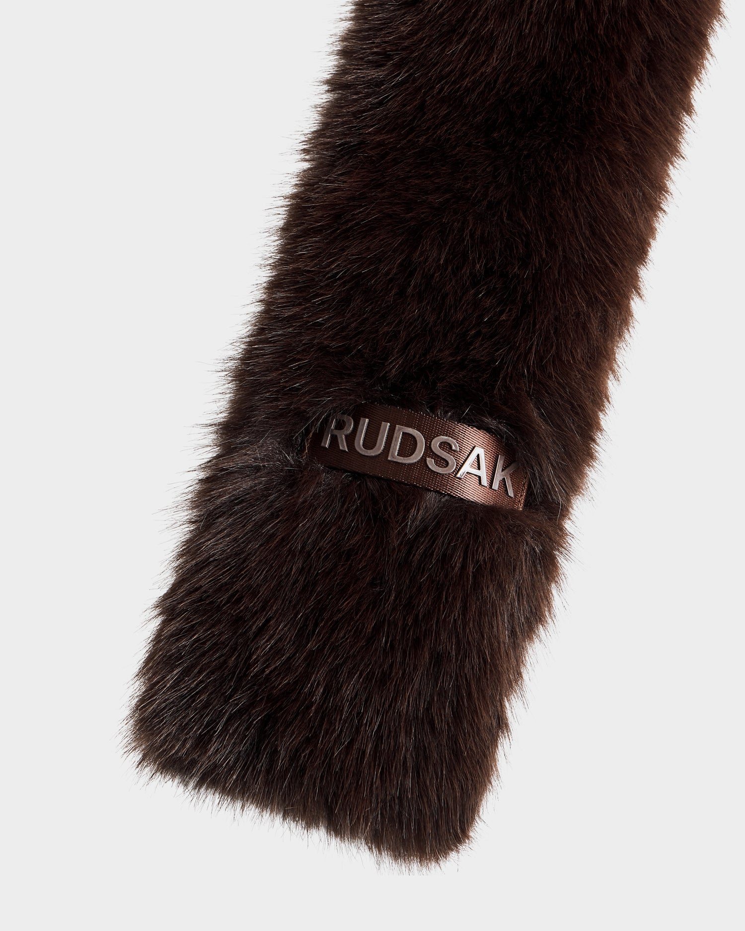 A close-up of Rudsak’s CARMY - L425534 Espresso, a women’s dark brown faux-fur scarf with a leather tag embossed with the brand name in silver. The accessory is set against a plain light gray background.