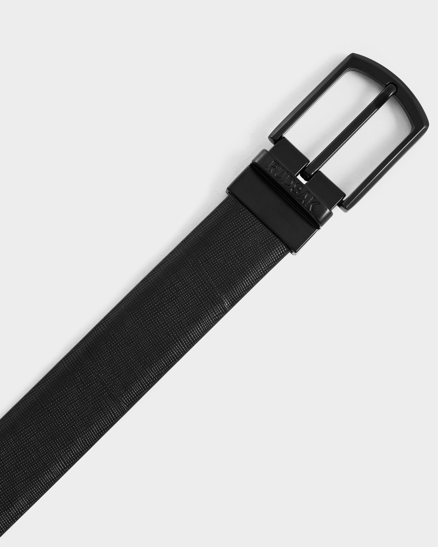 A close-up of the Rudsak RYLIE - M424500 Black reversible leather belt with a textured finish and shiny black buckle, elegantly displayed on a white background.