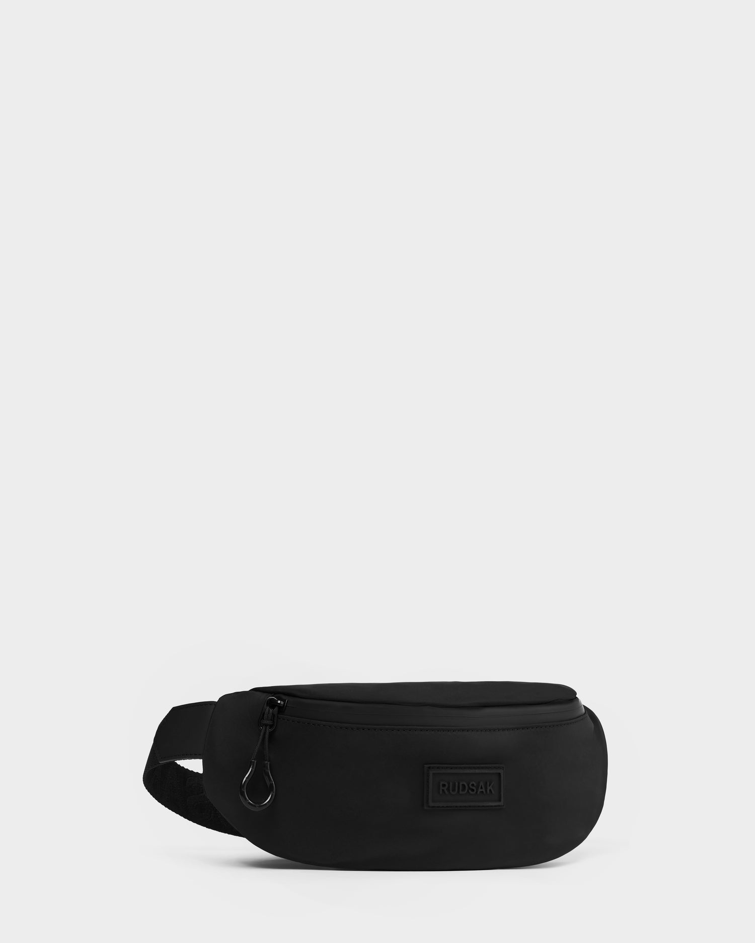 The Rudsak RILO - 9326000 Black waist bag features a zipper, adjustable strap, and a rectangular patch logo on the front, shown against a plain light background.