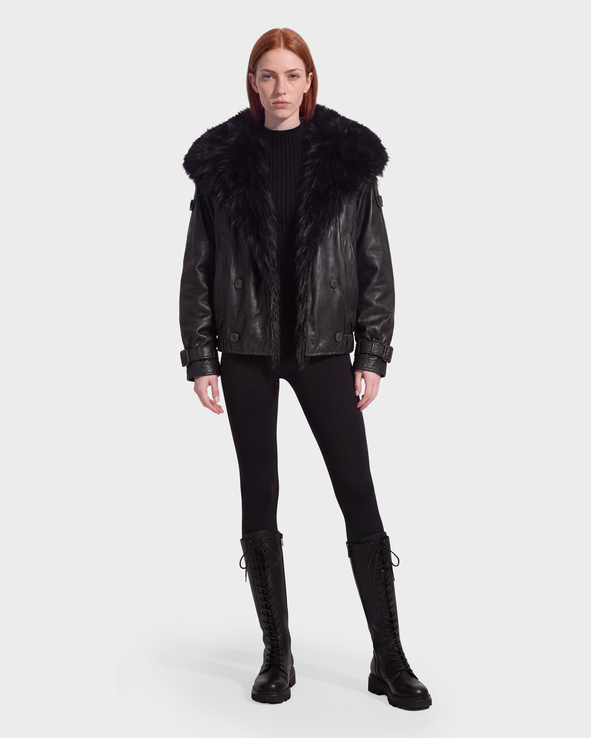 A woman wears a Rudsak JANELLE - 8126936 Black leather bomber with a faux fur collar over a black sweater and leggings, paired with black lace-up knee-high boots to create a modern silhouette.