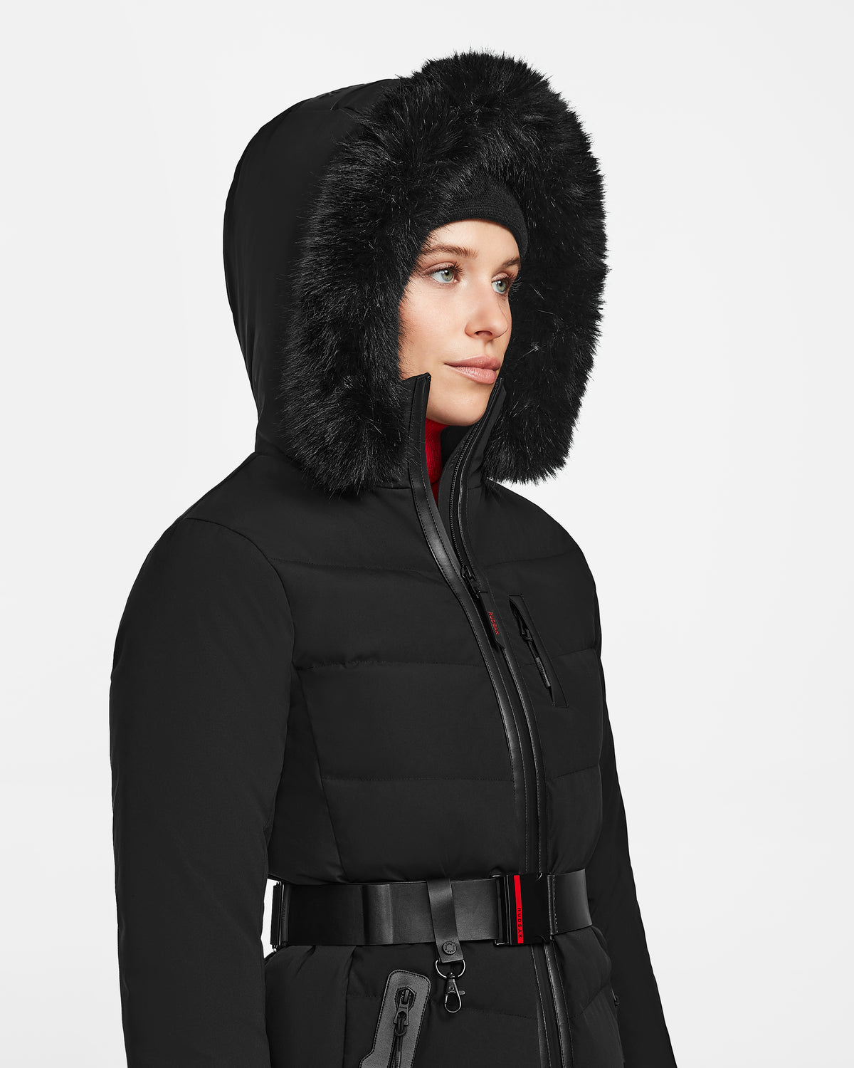 A woman wearing the Rudsak ALAIA - 8124816 Black quilted ski jacket with a fur-lined hood, front zipper, zippered pockets, belted waist, and a black beanie looks to the side.