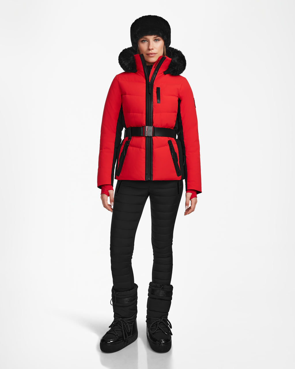 A woman wearing the Rudsak ALAIA - 8124816 Flame Red fitted ski jacket stands facing forward against a white background, paired with black pants, black fur-lined boots, and a black fur hat.