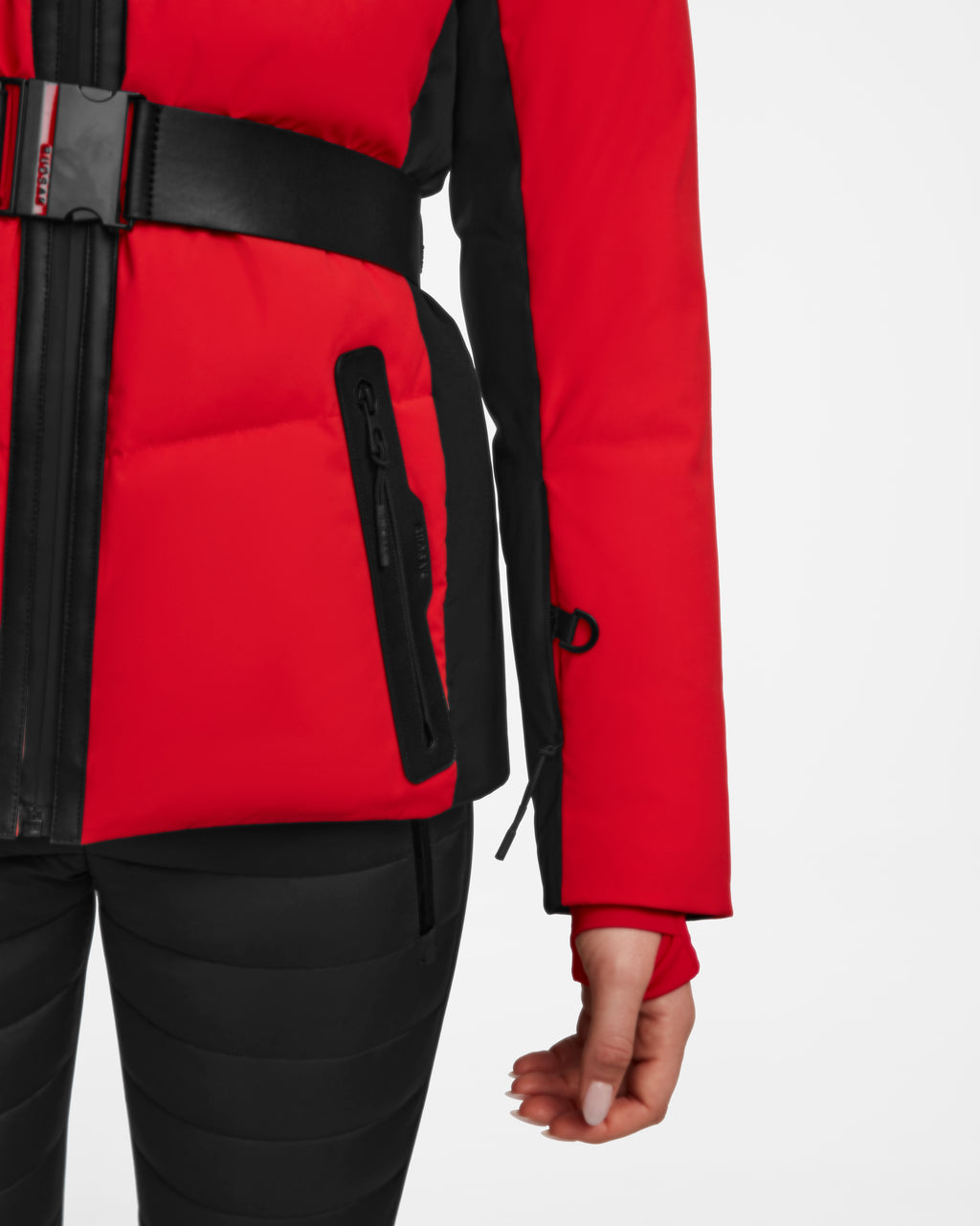 A person wears the Rudsak ALAIA - 8124816 Flame Red womens quilted ski jacket, featuring a belt and zippered pocket, paired with black pants. Only the lower half of the fitted jacket and part of one arm and hand are visible.