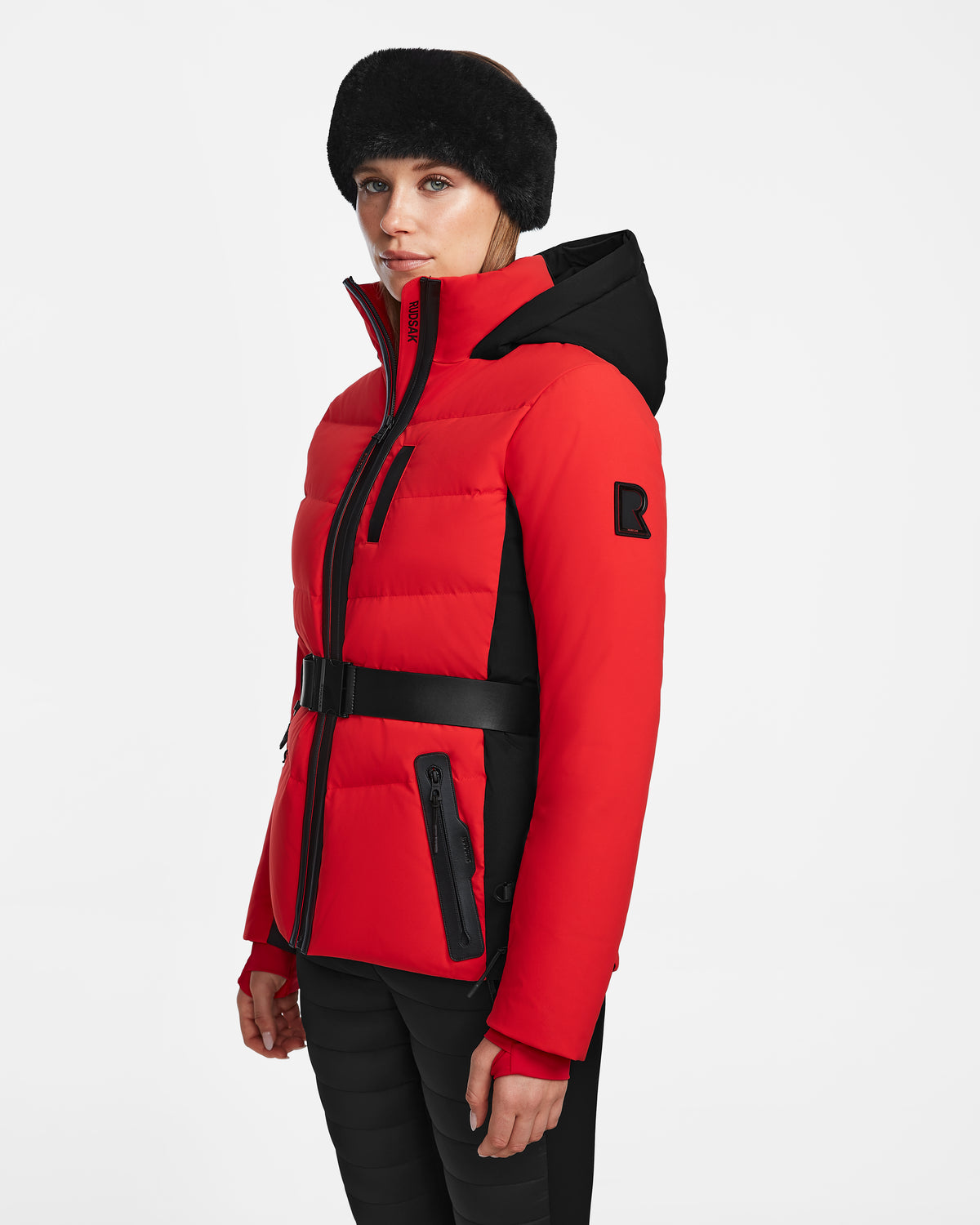 A woman in the Rudsak ALAIA - 8124816 Flame Red fitted quilted ski jacket with a red and black belt and black fur hat stands against a plain white background, facing slightly to the side with a neutral expression.
