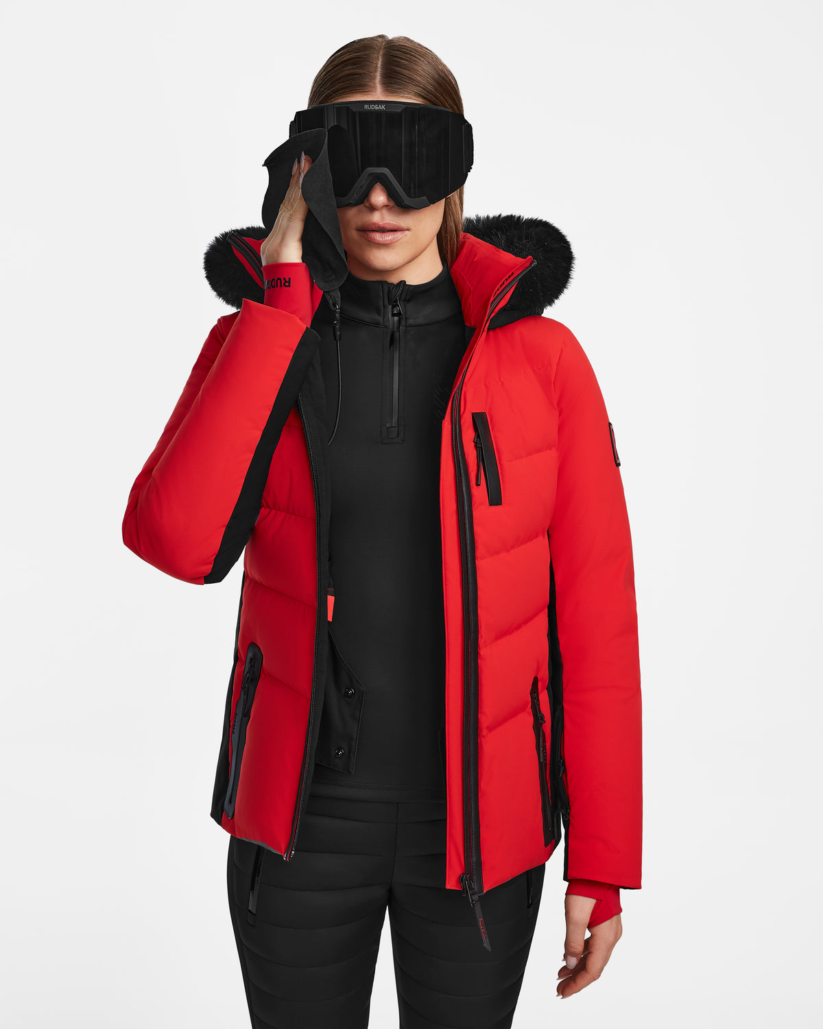 A woman adjusts large black ski goggles while wearing the Rudsak ALAIA - 8124816 Flame Red womens quilted ski jacket with black accents and a fur-lined hood, paired with black pants and a top, against a plain white background.