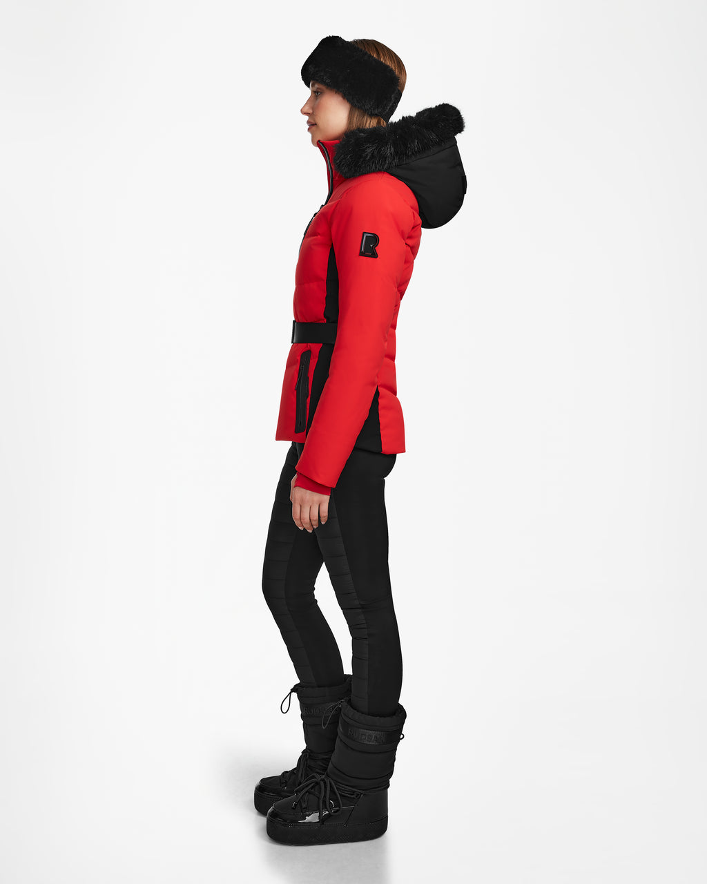 A person in profile wears the Rudsak ALAIA - 8124816 Flame Red womens quilted ski jacket with a fur-trimmed hood, paired with black leggings, winter boots, and a black headband against a plain white background.