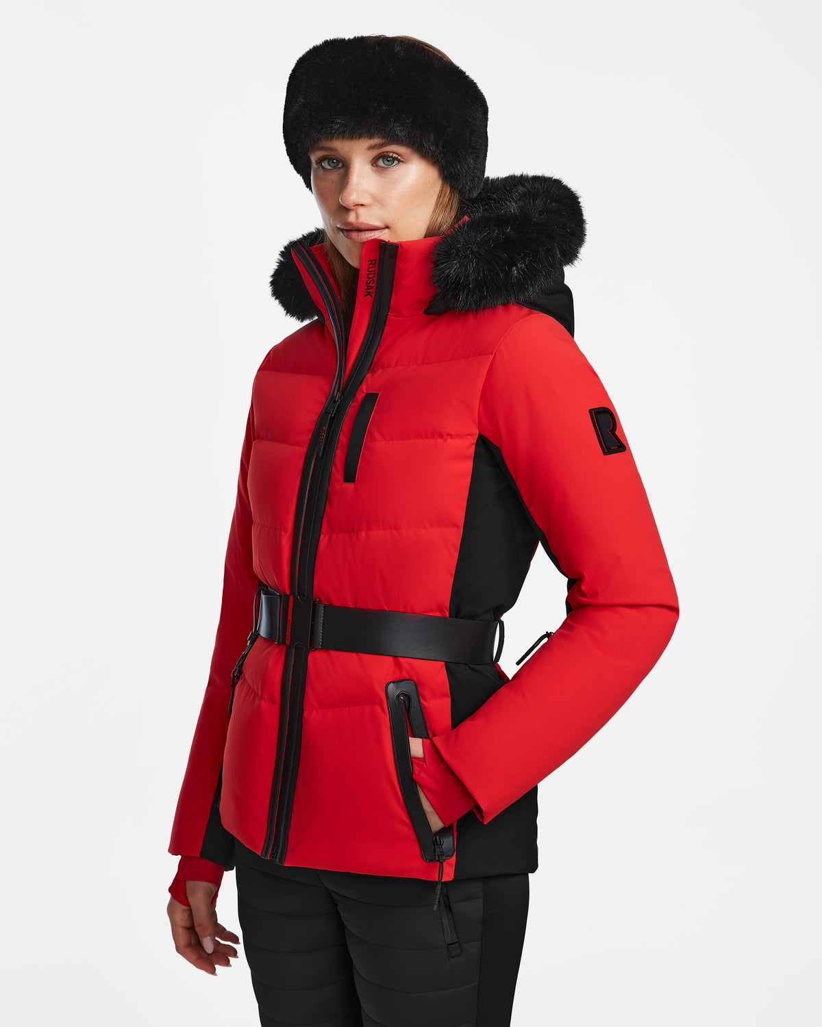 A woman models the Rudsak ALAIA - 8124816 Flame Red womens ski jacket with a fur-lined hood and wide belt, paired with a black fur hat, standing slightly left against a plain white background.