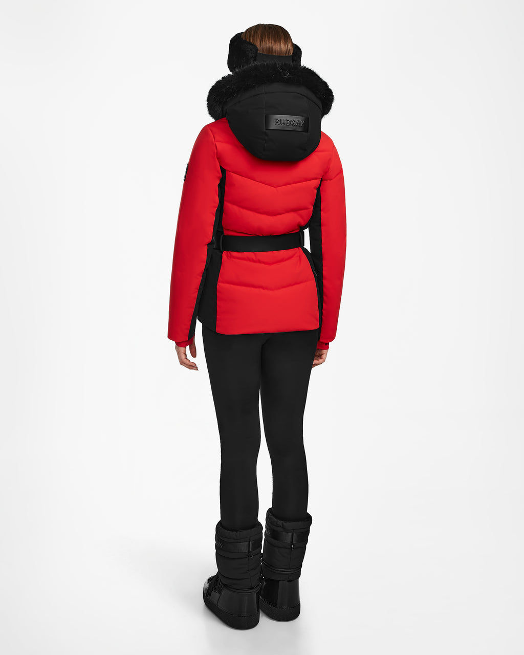 A person faces away, wearing the Rudsak ALAIA - 8124816 Flame Red womens quilted ski jacket with a fur-lined hood, black leggings, and black snow boots against a plain white background.