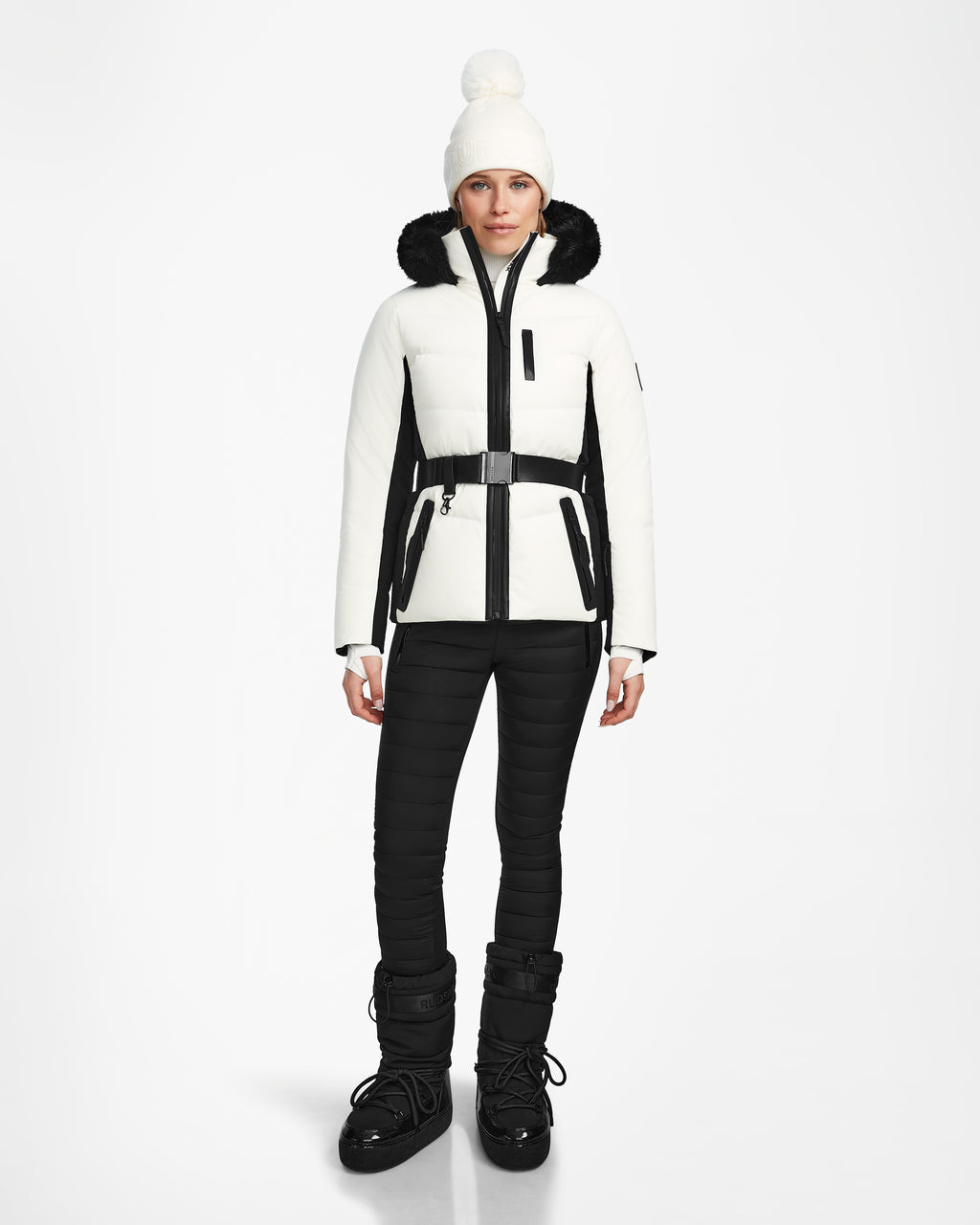 A woman stands facing forward wearing the Rudsak ALAIA - 8124816 Iceland ski jacket with black accents, black pants, black snow boots, and a white knit beanie with a pom-pom against a plain white background.