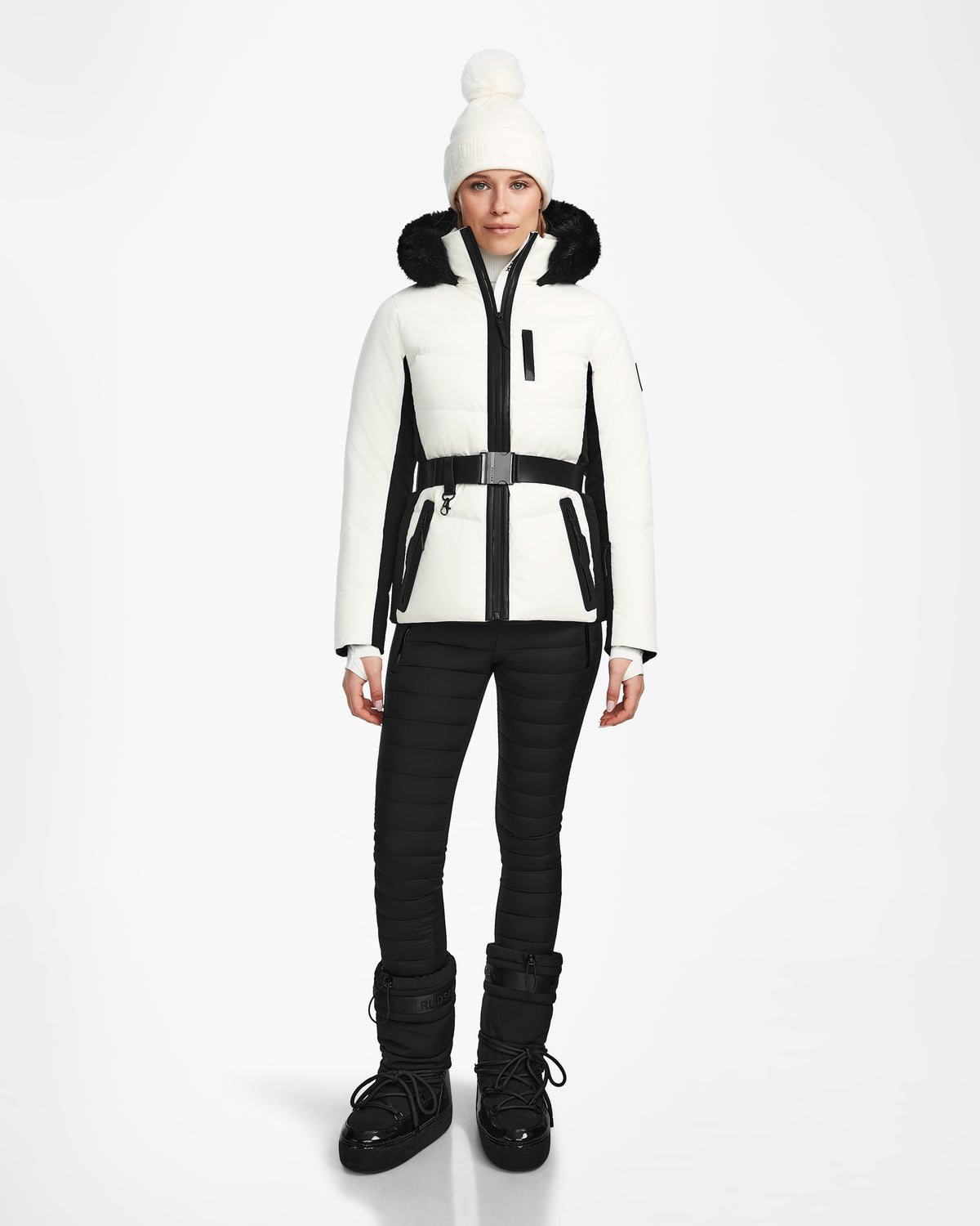 A woman stands facing forward wearing the Rudsak ALAIA - 8124816 Iceland ski jacket with black accents, black pants, black snow boots, and a white knit beanie with a pom-pom against a plain white background.