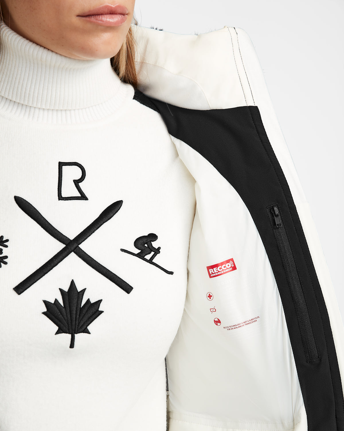A woman in a white turtleneck with black ski-inspired embroidery opens the Rudsak ALAIA - 8124816 Iceland womens quilted ski jacket, revealing a RECCO label on the inside. Only her lower face and upper body are visible.