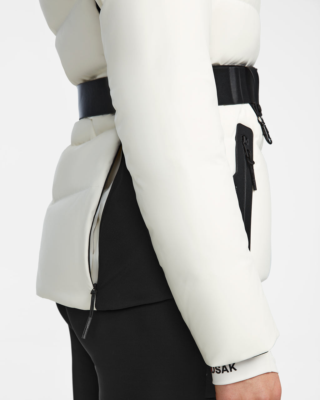 A close-up side view of the Rudsak ALAIA - 8124816 Iceland ski jacket in white with black accents, featuring a belt and zippered pocket. Only the midsection, part of the arm, and leg are shown.