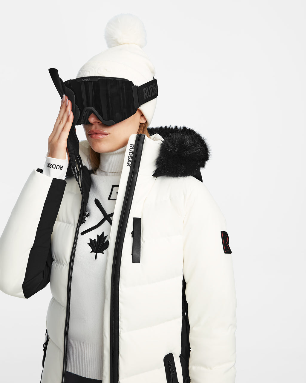 A person wearing the Rudsak ALAIA - 8124816 Iceland women’s quilted ski jacket with black accents, a white beanie, black ski goggles, and a maple leaf sweater adjusts their goggles against a white background.