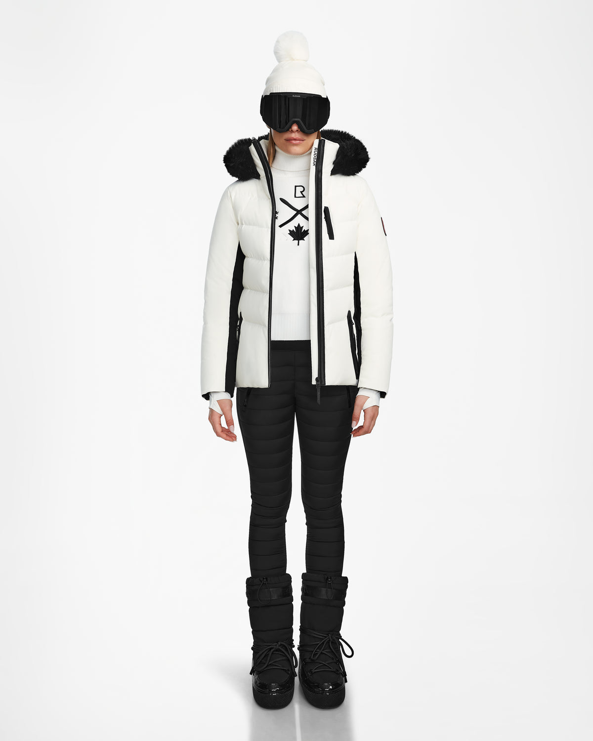 A person stands facing forward wearing the Rudsak ALAIA - 8124816 Iceland womens quilted ski jacket with black accents, black ski pants, snow boots, gloves, a white beanie with a pom-pom, and large black ski goggles.