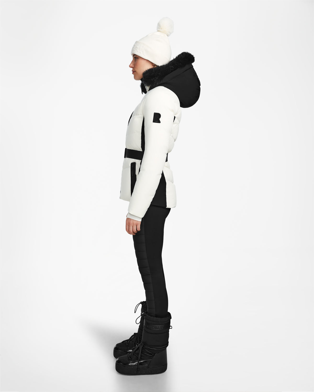 A person in profile wears the Rudsak ALAIA - 8124816 Iceland women’s quilted ski jacket with a black fur-lined hood, paired with black pants, winter boots, and a white pompom beanie, standing against a plain white background.