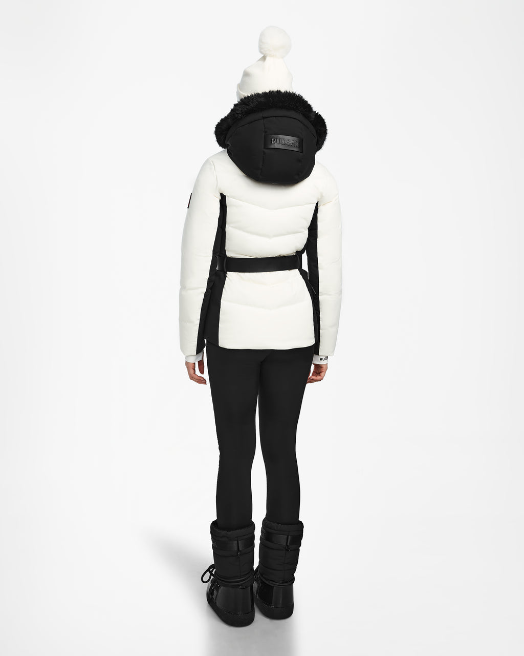 A person faces away, wearing the Rudsak ALAIA - 8124816 Iceland womens quilted ski jacket with a black hood, paired with black leggings, winter boots, and a white pom-pom beanie against a plain white background.