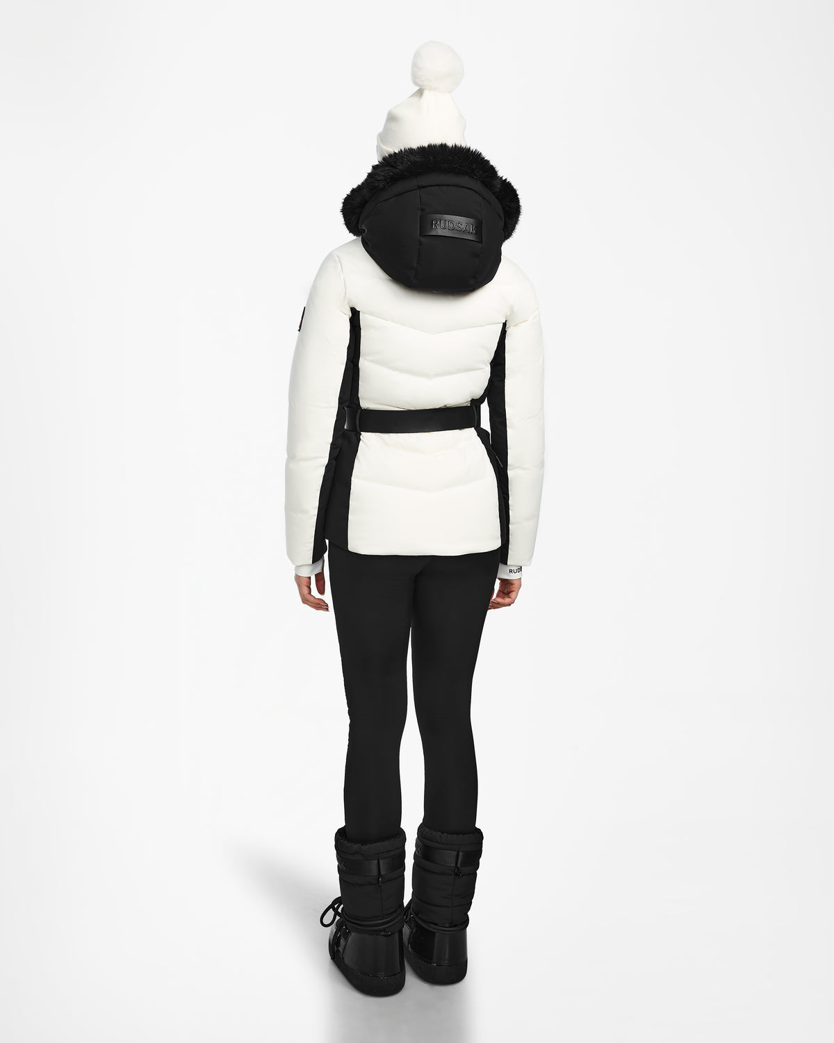A person faces away, wearing the Rudsak ALAIA - 8124816 Iceland womens quilted ski jacket with a black hood, paired with black leggings, winter boots, and a white pom-pom beanie against a plain white background.