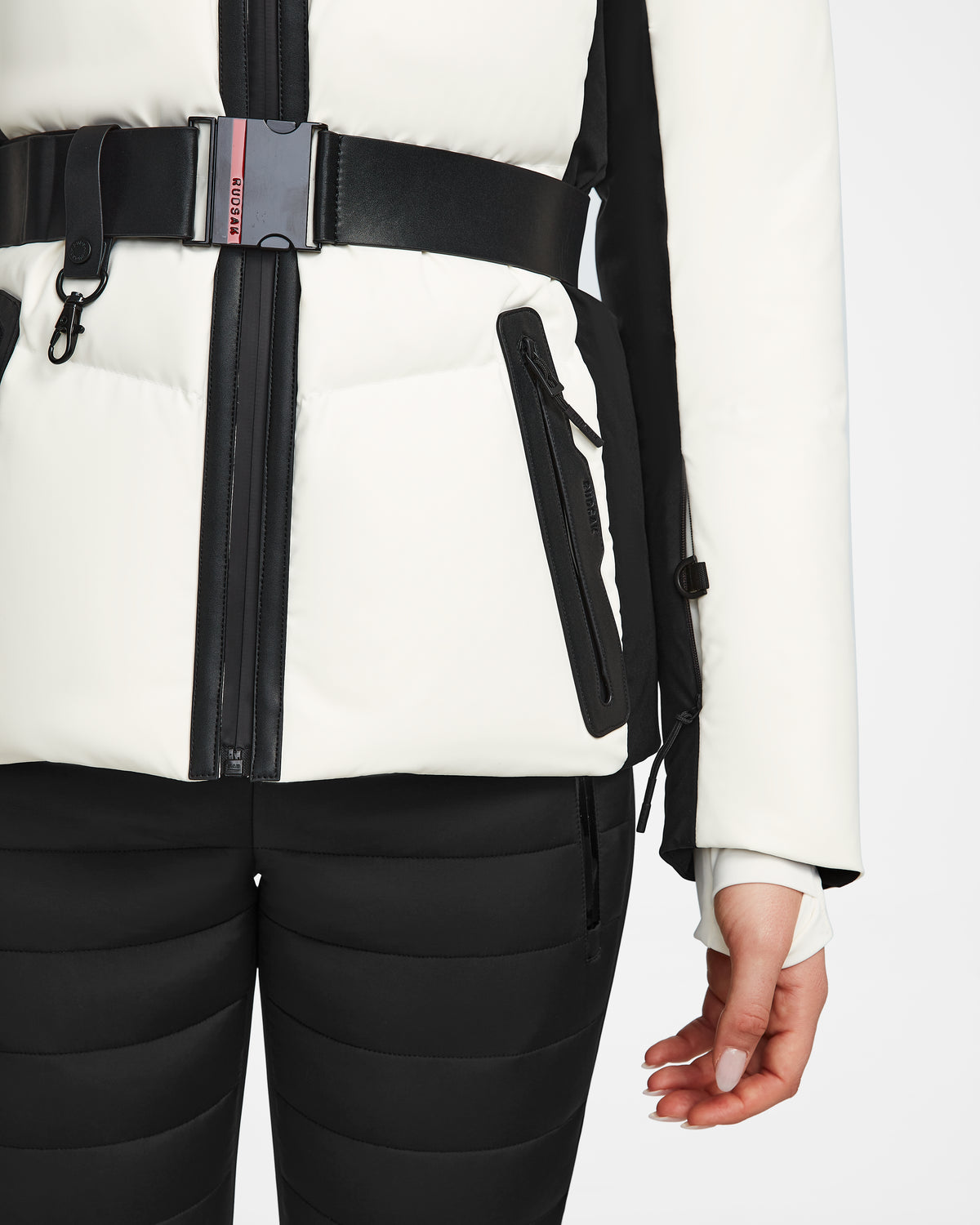 A person wearing the Rudsak ALAIA - 8124816 Iceland, a high-performance ski jacket in white with black accents and a belt, paired with black quilted pants. The jacket has zippered pockets, a front zipper, and their right hand is visible.