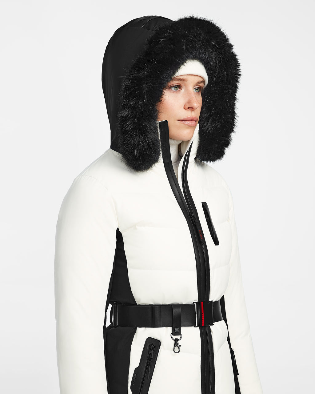A person wears a Rudsak ALAIA - 8124816 Iceland womens quilted ski jacket with a fur-lined hood, black belt, and white beanie, facing slightly to the right against a plain background.