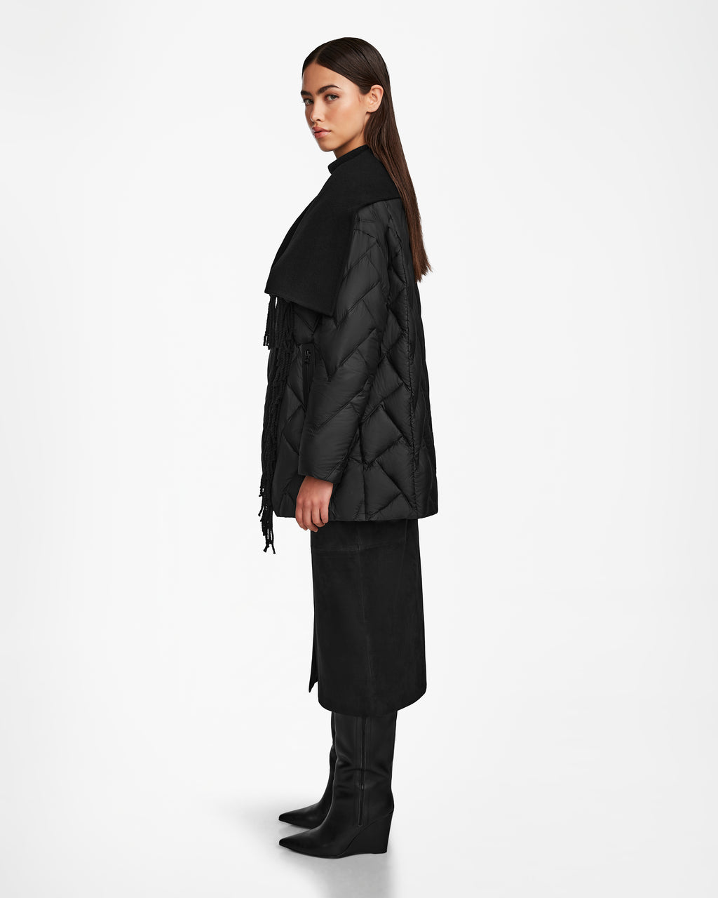 A woman stands in profile against a white backdrop wearing the ANNE MARIE - 8124713 Black quilted down wool jacket by Rudsak, styled with a large scarf, black midi skirt, and knee-high boots. Her straight hair is tucked behind her ears.