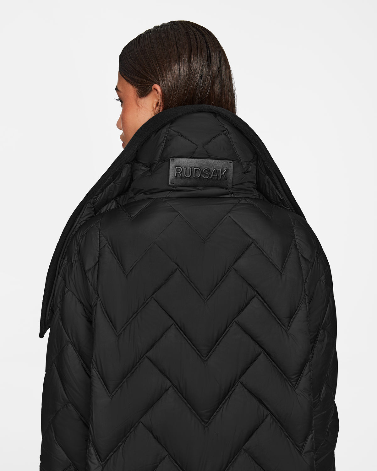 A person with straight brown hair wears the Rudsak ANNE MARIE - 8124713 black mid-length women’s down wool jacket with a chevron pattern and leather patch, collar turned up, channeling the western trend against a white background.