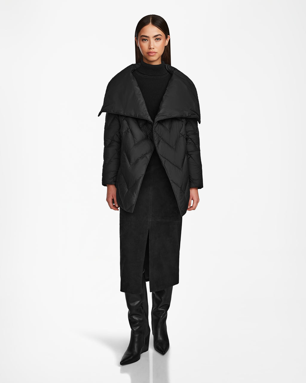 A woman stands facing forward in the Rudsak ANNE MARIE - 8124713 black quilted down wool jacket with a wide collar, styled with a black turtleneck, a front-slit midi skirt, and knee-high boots against a plain white background.