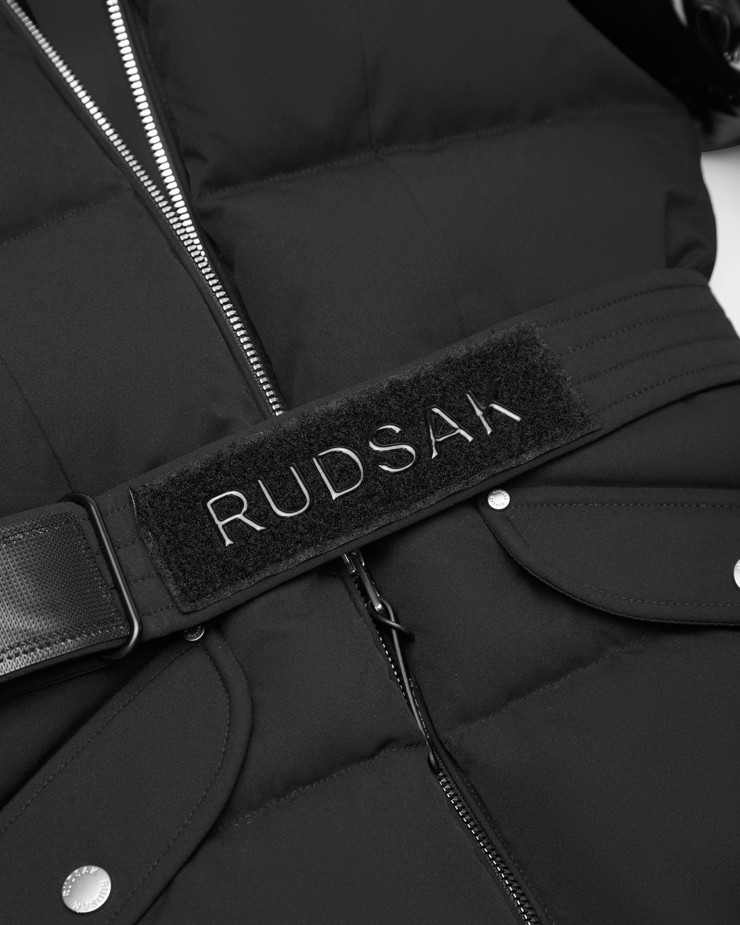 Close-up of the Rudsak AUDREY K - 0124504 Black girls' puffer jacket, made with RDS-certified down. This wind-resistant jacket features a zipper, snap-button pockets, and a belted Velcro patch embroidered with “RUDSAK” in white.