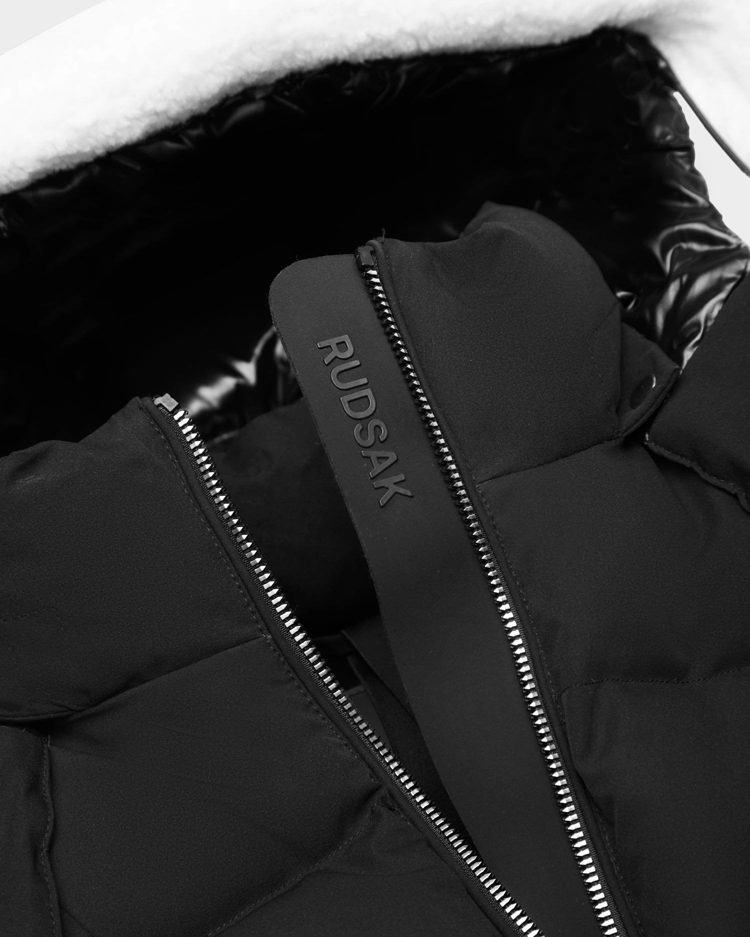 Close-up of the Rudsak AUDREY K - 0124504 Black girls' down puffer jacket with a silver zipper and RUDSAK tag. Features wind resistance, shiny black inner lining, RDS-certified down, and a white fluffy hood lining.