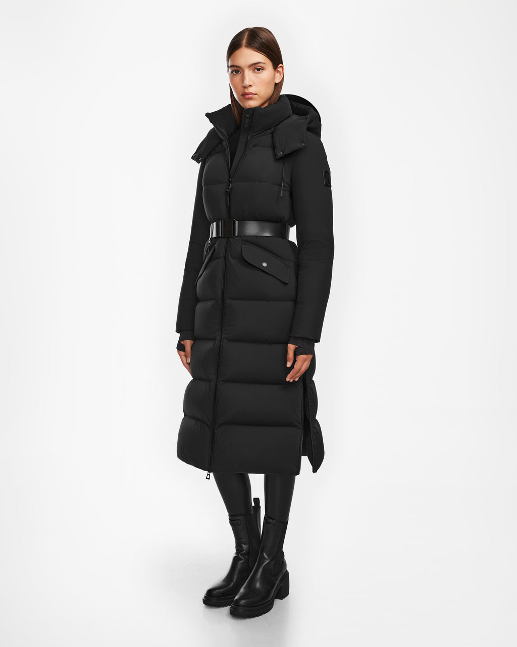A woman faces forward wearing the Rudsak CALI - 8124600 Black womens long down puffer winter jacket with high collar and hood, black leggings, and black ankle boots. This coat uses RDS-certified down for warmth and comfort.