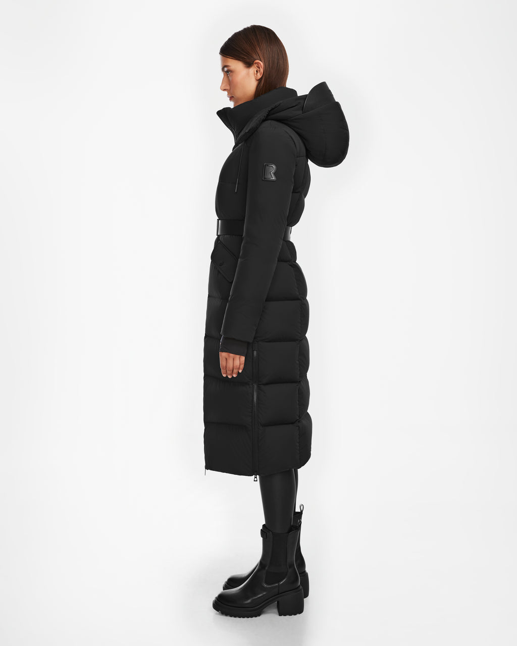 A woman in profile models the Rudsak CALI - 8124600 Black women’s long down puffer coat with a hood, paired with black leggings and chunky black boots against a white background.
