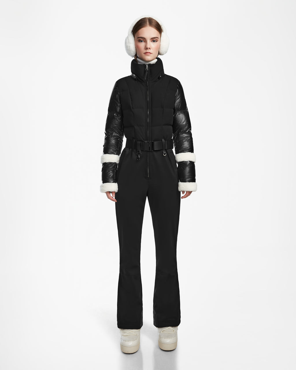 A woman stands against a white background wearing Rudsak’s CARMEN - 8124783 Black fitted one-piece ski suit, styled with shiny sleeves, white cuffs, white earmuffs, and beige platform snow boots featuring faux shearling details.