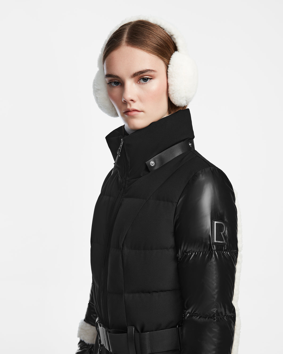 A young woman with straight brown hair wears the Rudsak CARMEN - 8124783 Black quilted winter coat with faux shearling and a belted waist, paired with white earmuffs, standing against a plain white background.