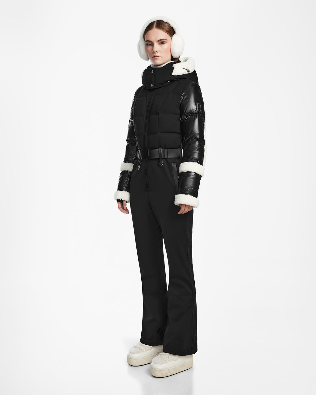 A woman stands against a white background wearing the Rudsak CARMEN - 8124783 Black puffer jacket with faux shearling details, styled with black belted pants, white earmuffs, and white platform shoes. She looks ahead with a neutral expression.