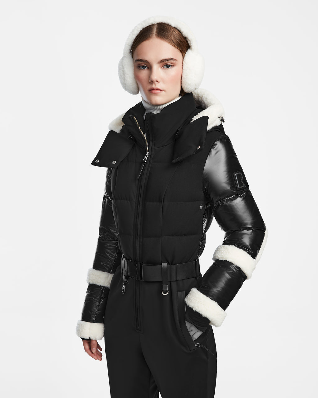 A woman wears the Rudsak CARMEN - 8124783 Black puffer jacket with white fur trim and RECCO rescue technology, paired with black belted pants, a white turtleneck, and white earmuffs against a plain white background.