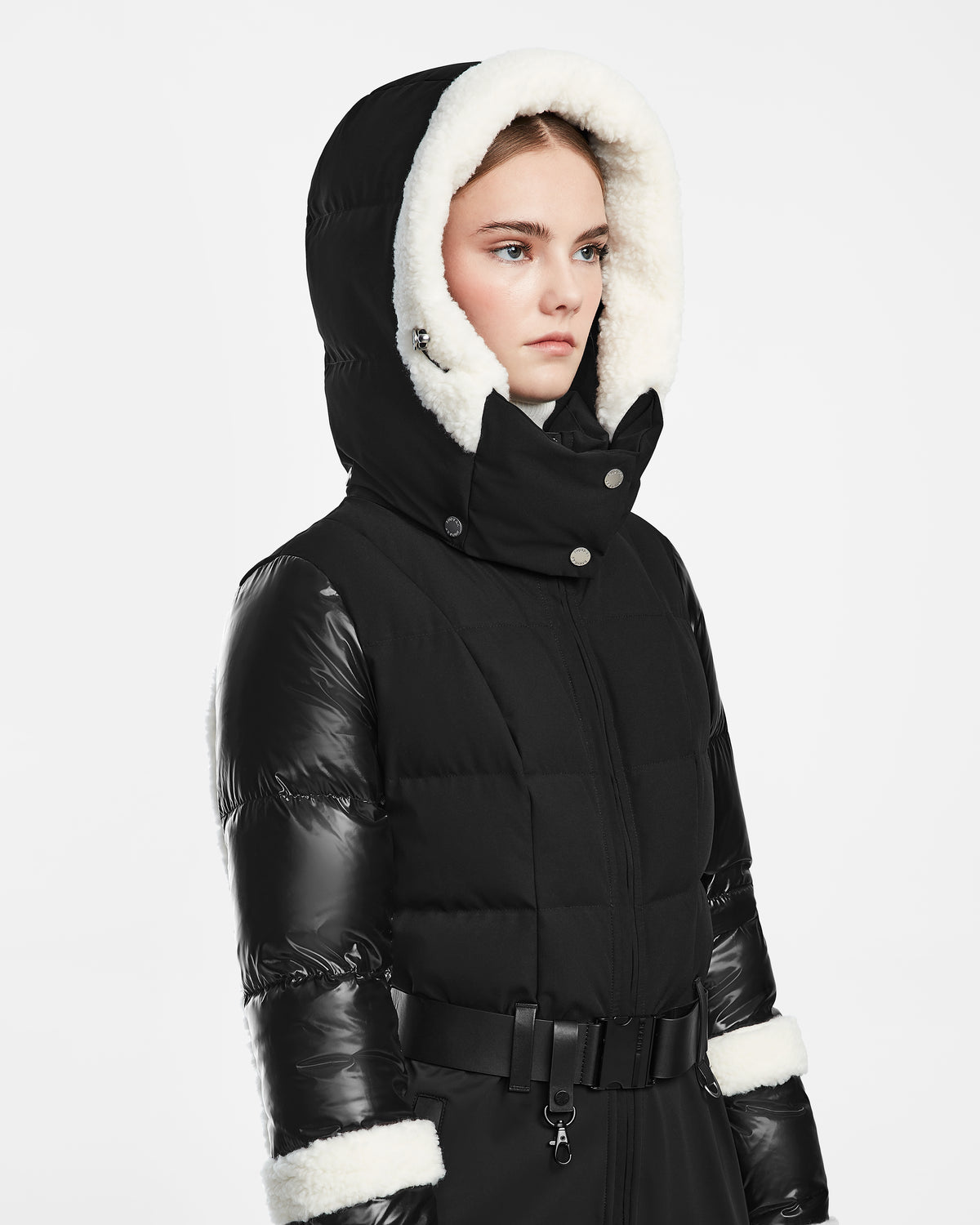 A young woman wears the Rudsak CARMEN - 8124783 Black puffer coat, featuring a belted waist, large white faux fur hood, and RECCO rescue technology, looking to the side against a plain white background.
