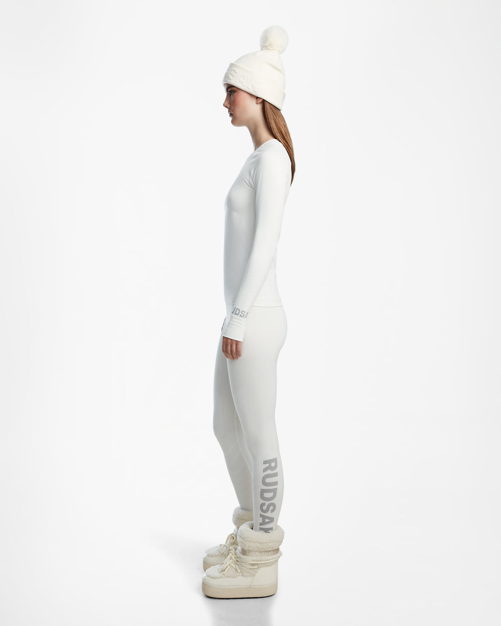 A woman stands in profile against a white background, wearing the Rudsak CARRIE - 8524619 Iceland knit hat with pom-pom, a long-sleeve women’s thermal top, stretch leggings with RUDSAK branding, and chunky white boots.
