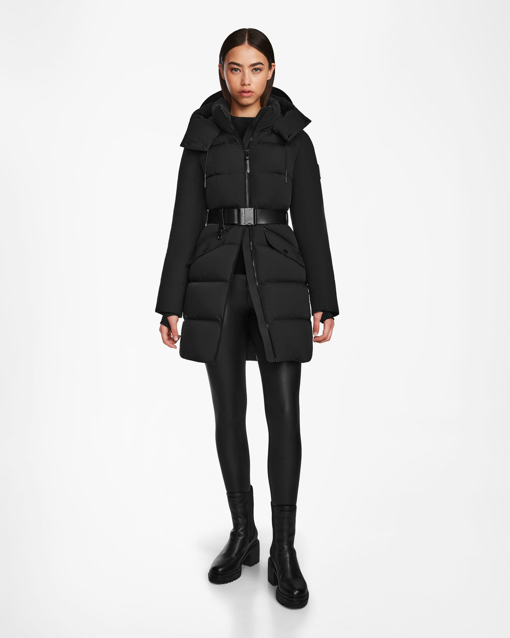 A woman stands against a white background, wearing the Rudsak CAT - 8124584 Black mid-length puffer coat with belt, paired with black leggings and boots. She faces forward neutrally, hands by her sides.