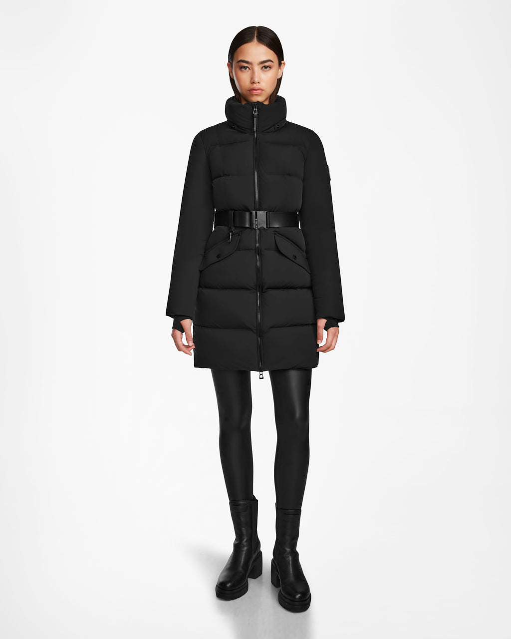 A woman faces forward wearing the Rudsak CAT - 8124584 Black mid-length puffer coat with a belt, paired with black leggings and boots, against a plain white background.