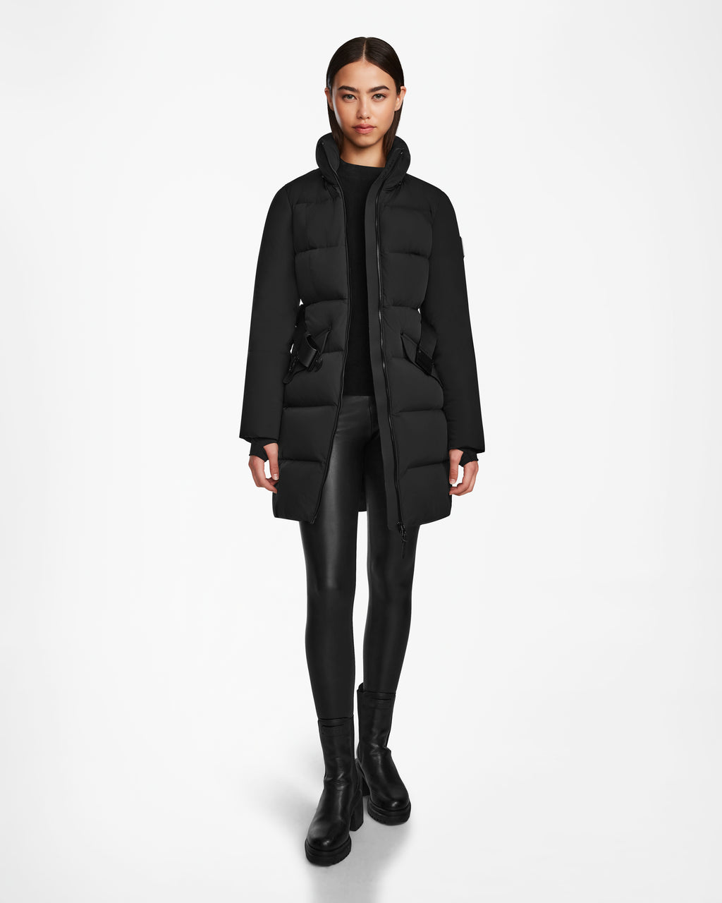 A woman with slicked-back hair and a neutral expression wears the Rudsak CAT - 8124584 Black womens quilted down puffer coat, styled with black leggings and ankle boots, standing against a plain white background.
