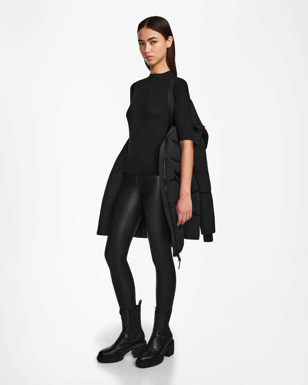 A woman stands against a white background wearing Rudsaks CAT - 8124584 Black quilted down puffer over a black fitted top, faux leather leggings, and black boots.