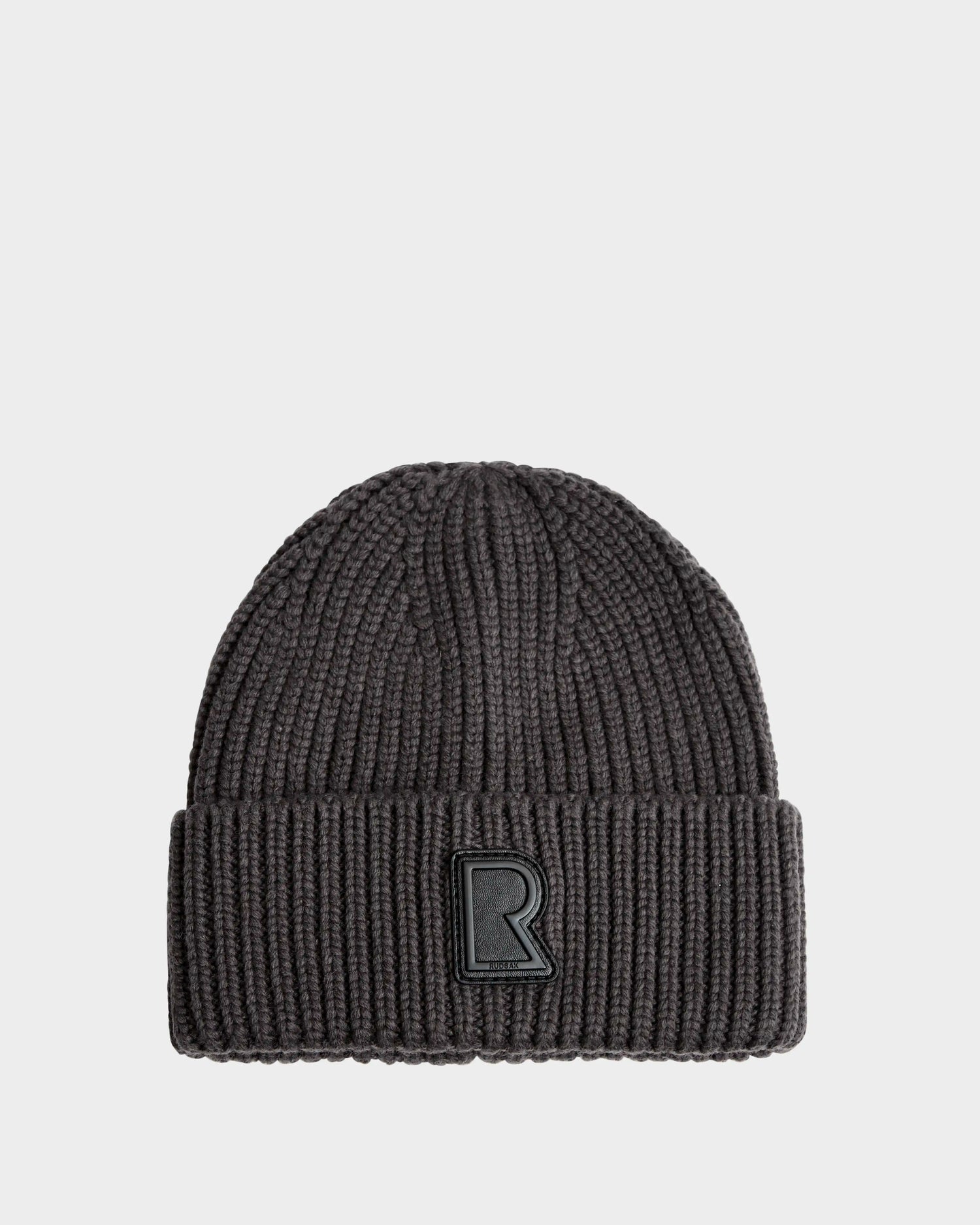 The CYBELLE - 9424504 Grey by Rudsak is a dark gray ribbed knit beanie with a folded brim and a black patch featuring the letter R on the front, shown on a plain white background.