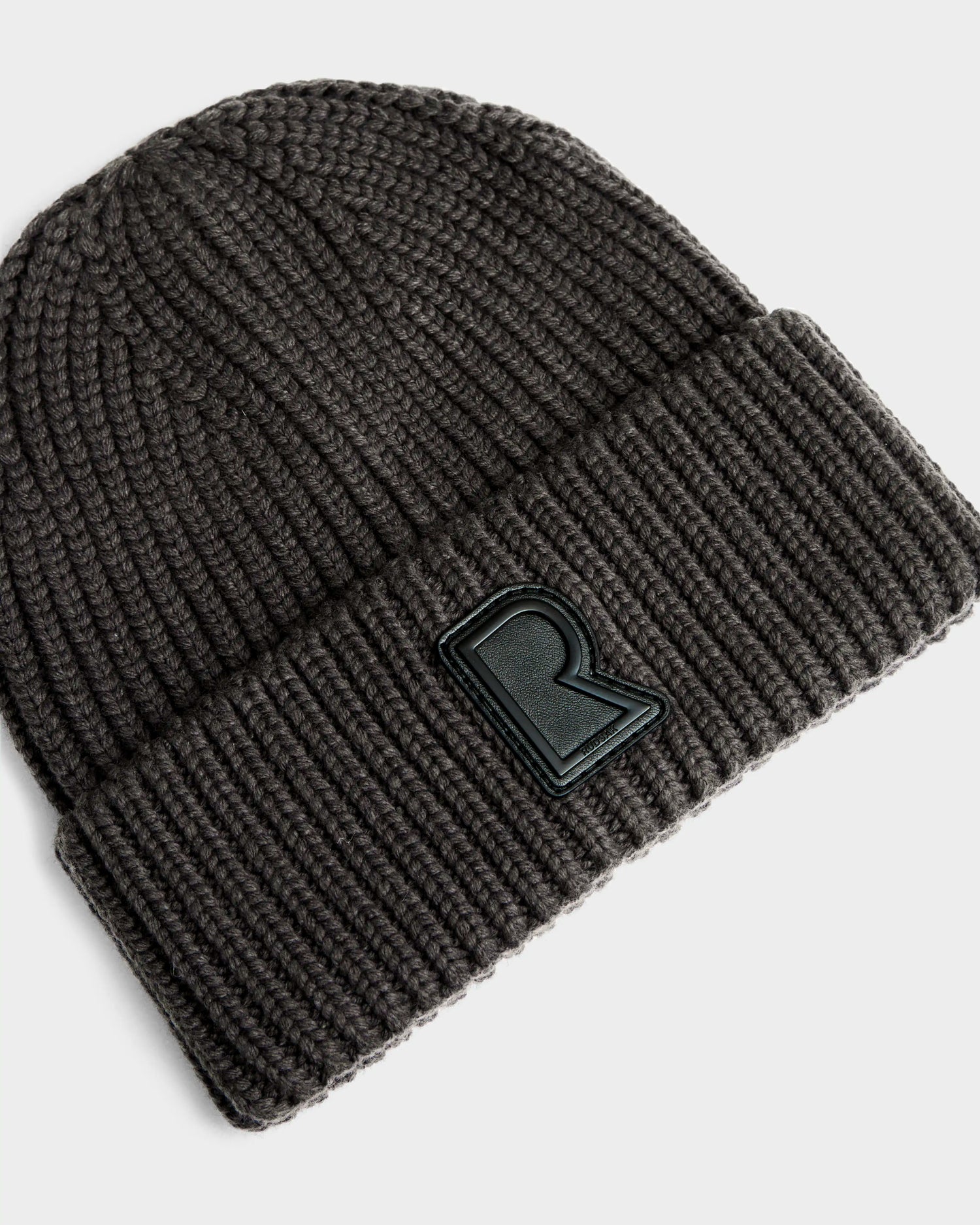 The Rudsak CYBELLE - 9424504 Grey is a unisex dark gray ribbed knit hat with a folded brim and a raised black R patch on the front—a stylish winter accessory.