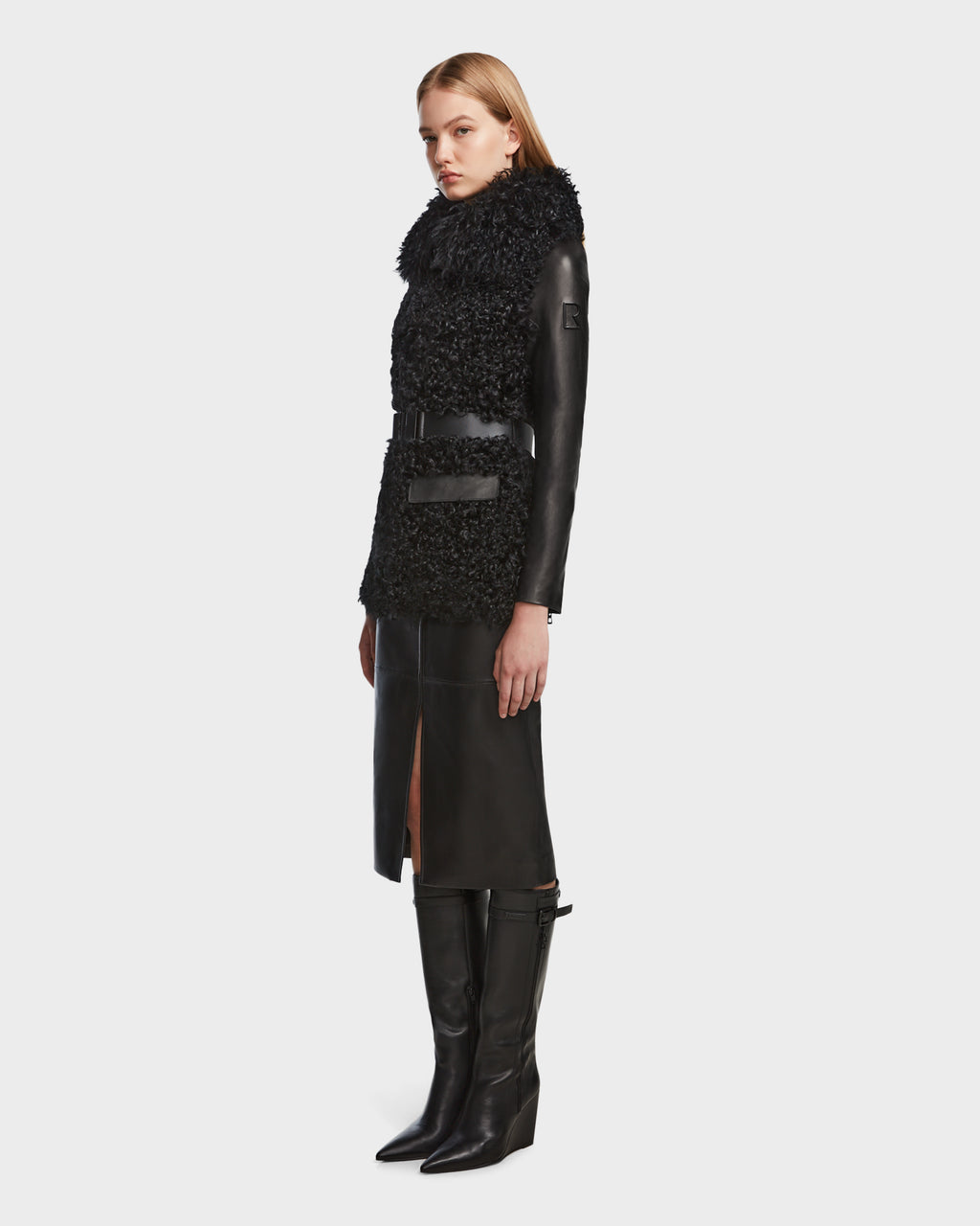 A woman stands in profile wearing the Rudsak DASHA - 8124809 Black jacket with a matching belt and knee-high black boots. She has straight blonde hair and a neutral expression against a plain light background.