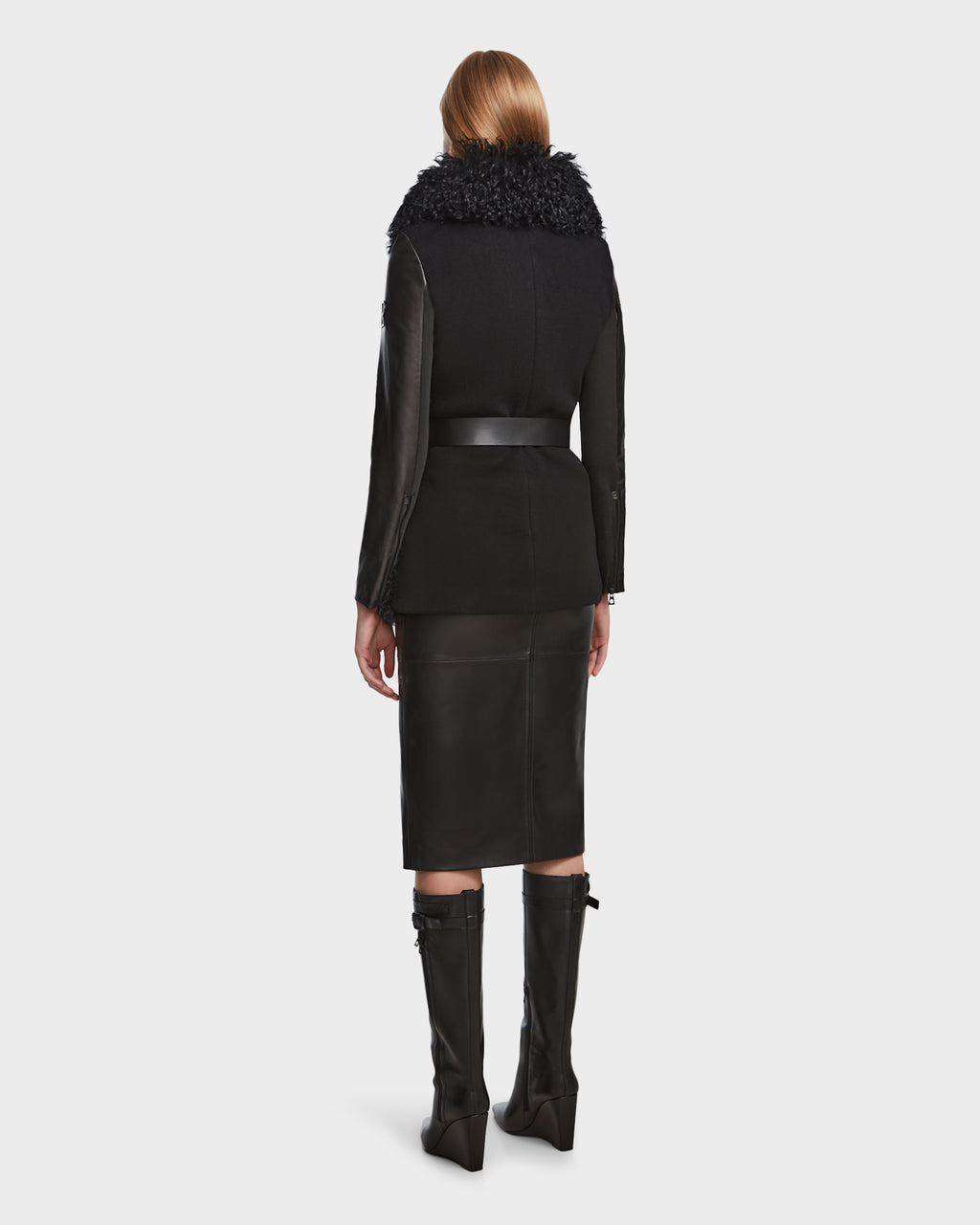 A woman with straight blonde hair is seen from behind, wearing the Rudsak DASHA - 8124809 Black womens leather shearling jacket with a textured fur collar, a black knee-length skirt, and knee-high black boots against a plain light background.
