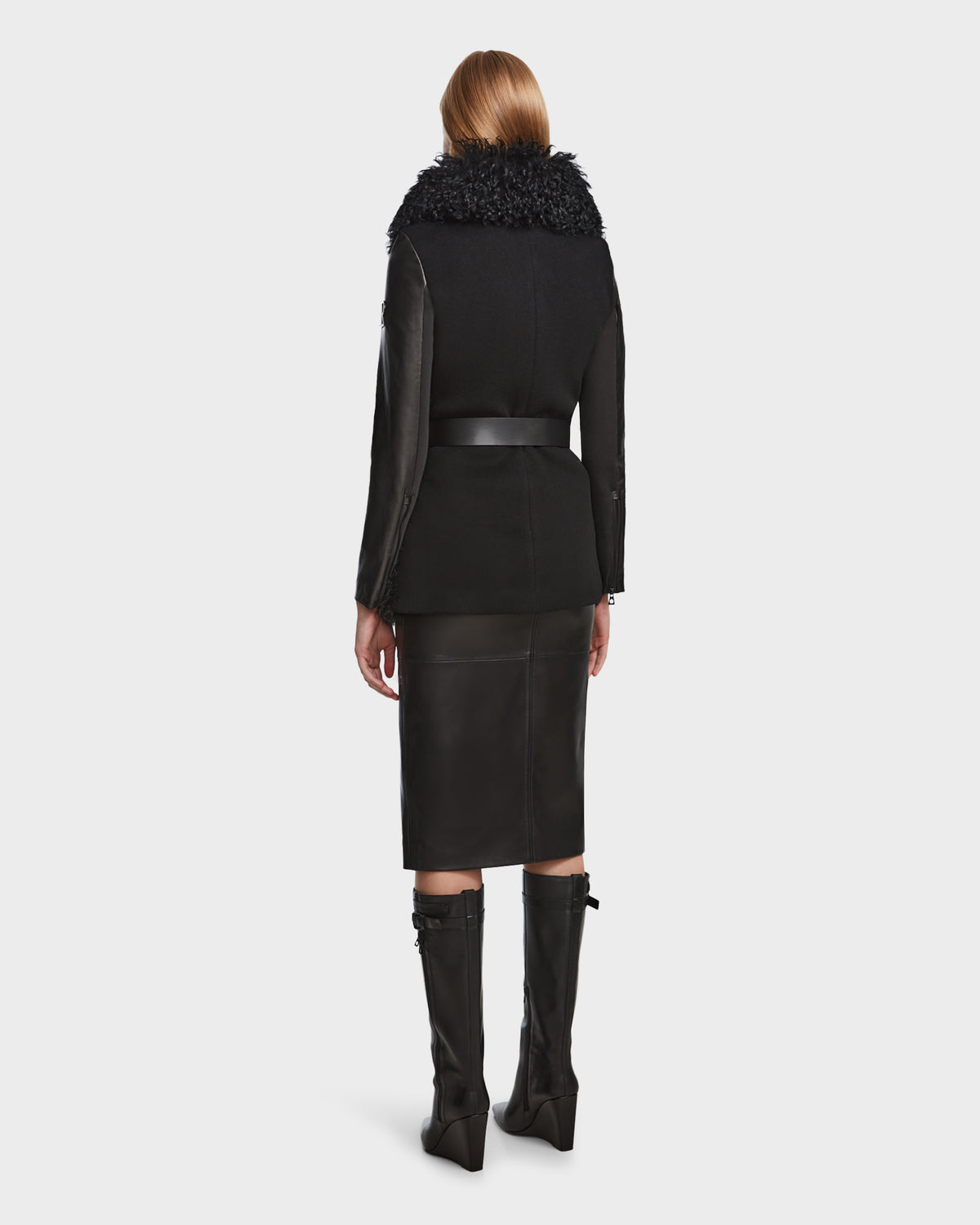 A woman with straight blonde hair is seen from behind, wearing the Rudsak DASHA - 8124809 Black womens leather shearling jacket with a textured fur collar, a black knee-length skirt, and knee-high black boots against a plain light background.