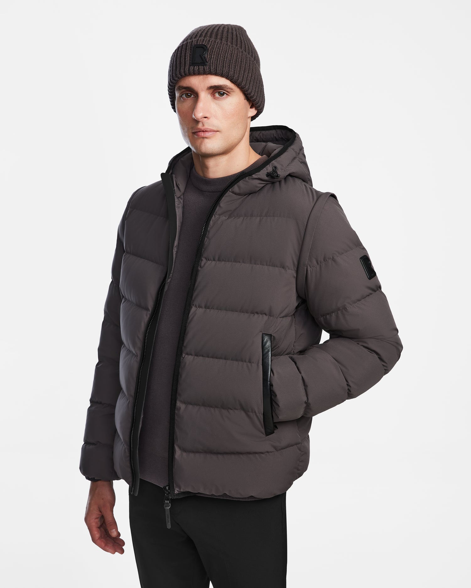 A man stands against a white background wearing the Rudsak DEMIAN M-R - M123504 Grey mens down puffer jacket with a hood and black zipper, paired with a dark knit beanie and black pants, looking straight at the camera.